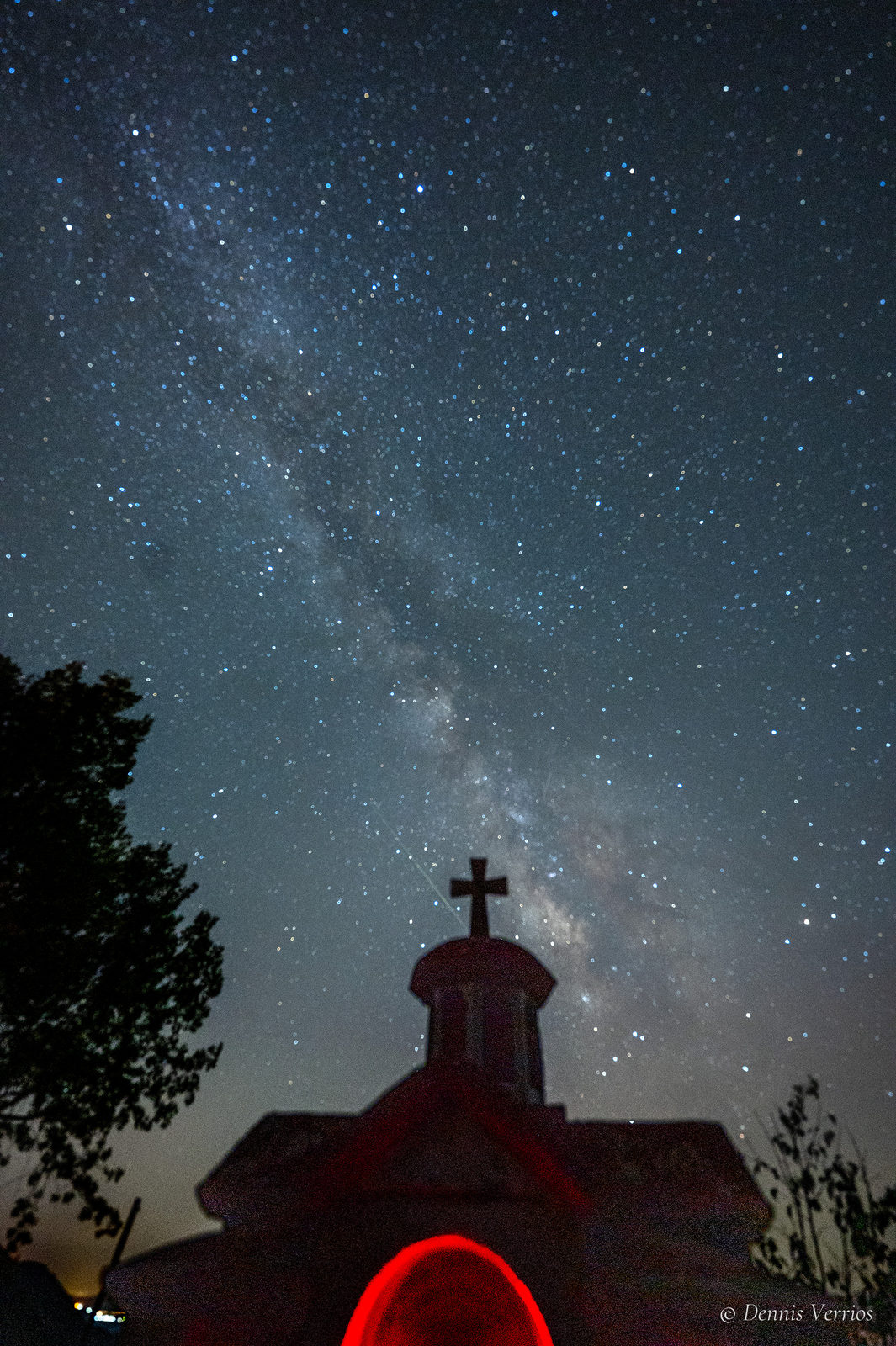A church bell tower topped with a cross, lit red from below, under the Milky Way and a sky full of stars in Dimitsana, Arcadia, Greece. Photograph by Dennis Verrios.