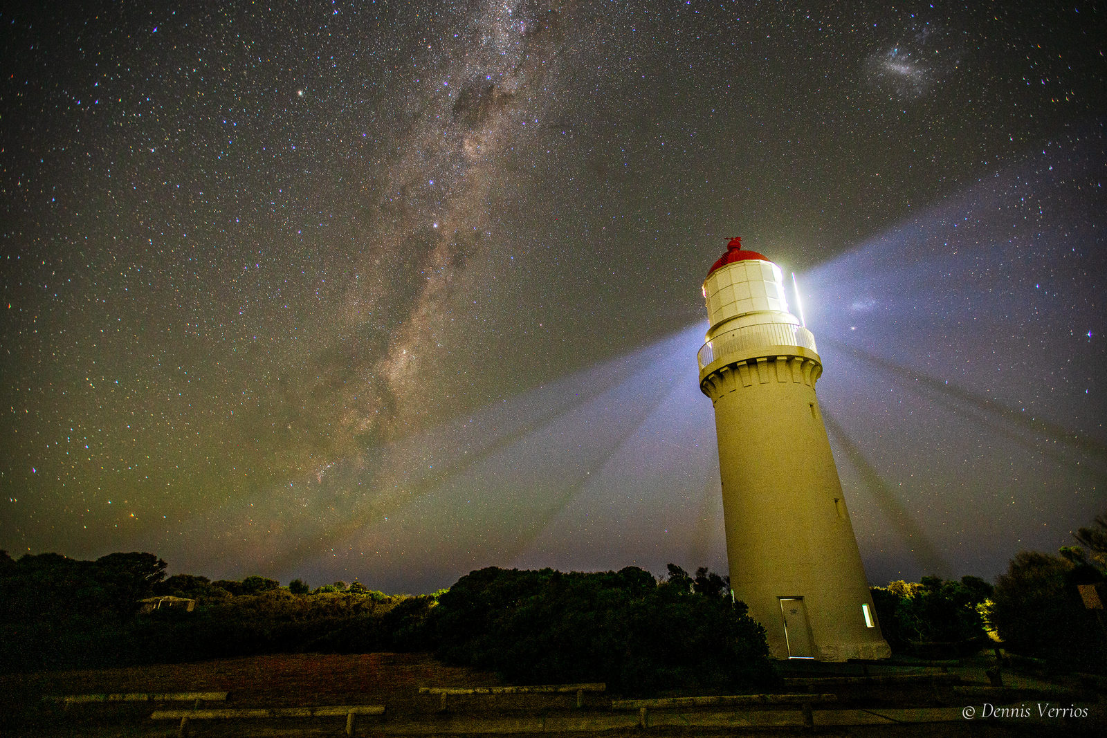 The band of the Milky Way stretching diagonally across a star-filled sky above the lit Cape Schanck Lighthouse and coastal scrub. Photograph by Dennis Verrios.