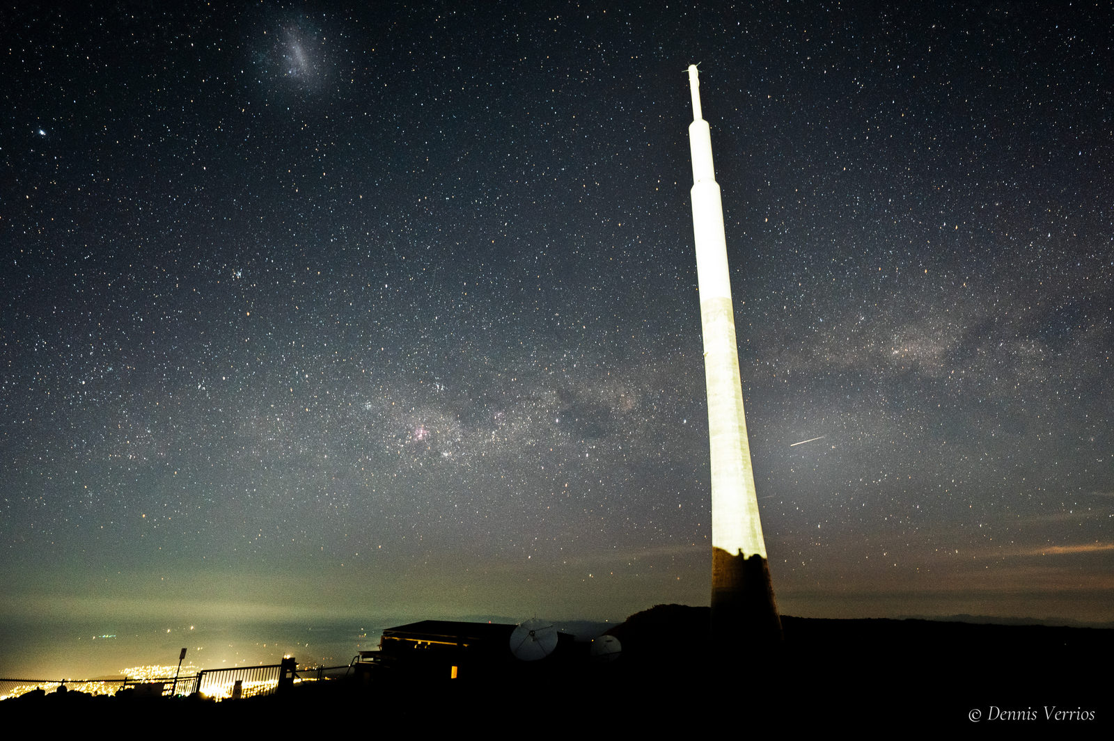 The lit broadcast tower on the summit of kunanyi / Mount Wellington under a starry sky, with the Large Magellanic Cloud at top left, the Milky Way across the middle, a meteor streak beside the tower and the lights of Hobart below. Photograph by Dennis Verrios.