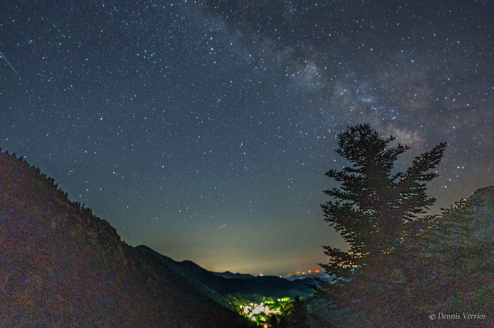 The Milky Way arching over a fir tree on a mountainside, with the lights of Alonistaina village glowing in the valley below, Arcadia, Greece. Photograph by Dennis Verrios.
