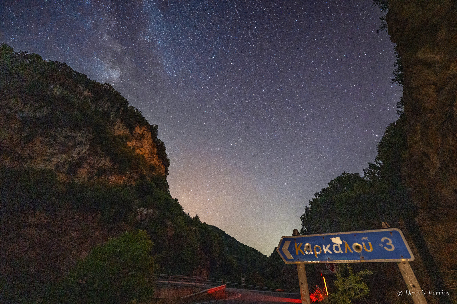 The Milky Way above a rocky gorge with a blue road sign reading Karkalou 3, in Arcadia, Greece. Photograph by Dennis Verrios.