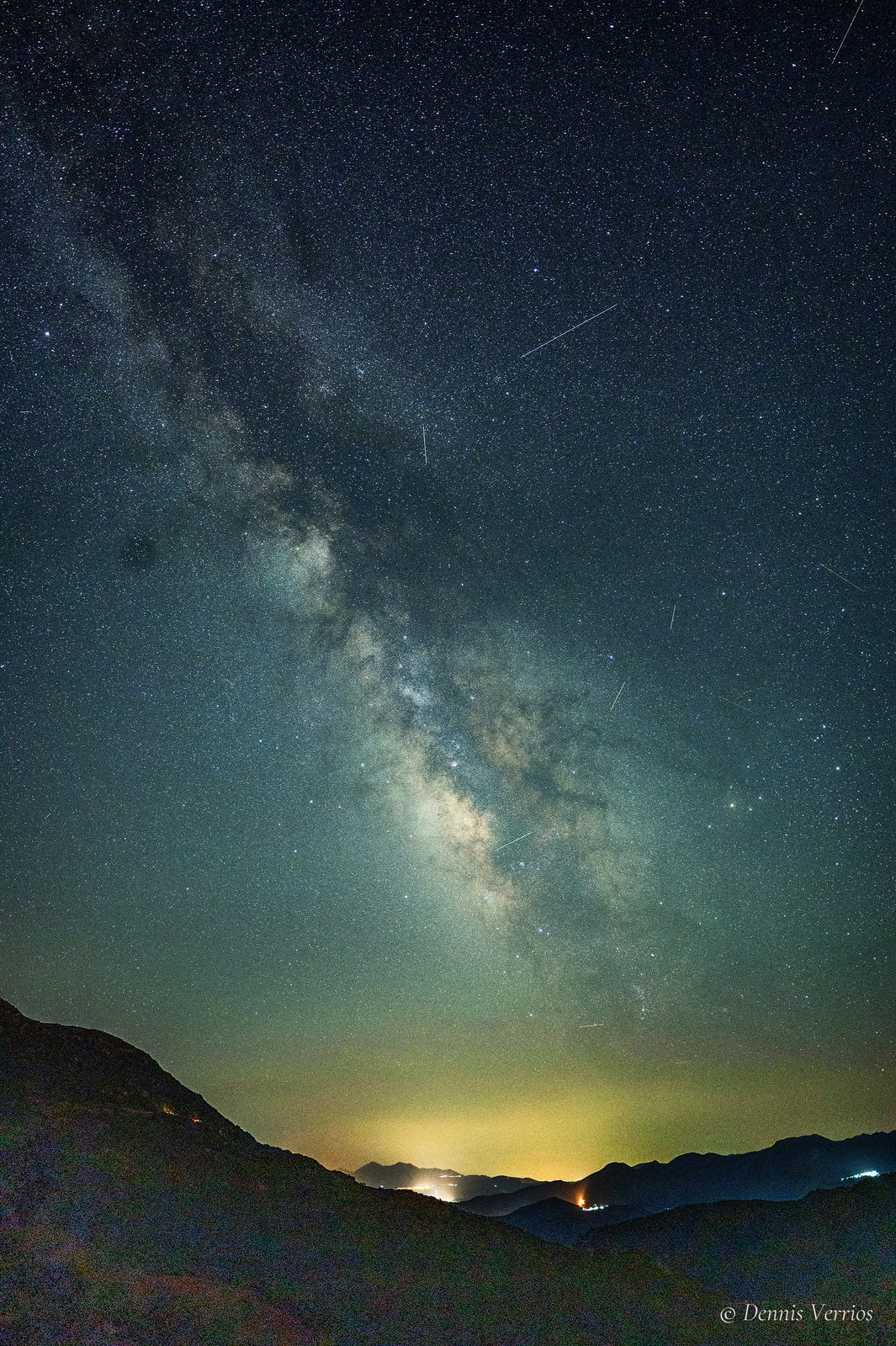 The Milky Way rising vertically above dark mountain ridges and the glowing lights of distant villages near Alonistaina, Arcadia, Greece, with a meteor streak in the sky. Photograph by Dennis Verrios.