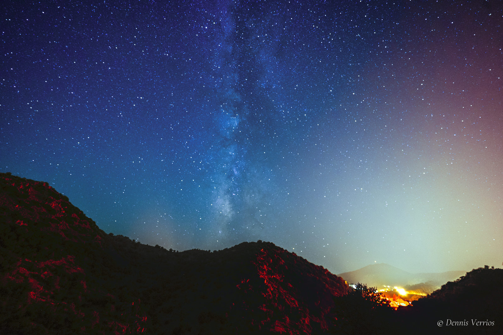 A vivid blue Milky Way rising above dark mountain ridges, with red-lit shrubs in the foreground and the orange glow of a town below, near Dimitsana, Arcadia, Greece. Photograph by Dennis Verrios.