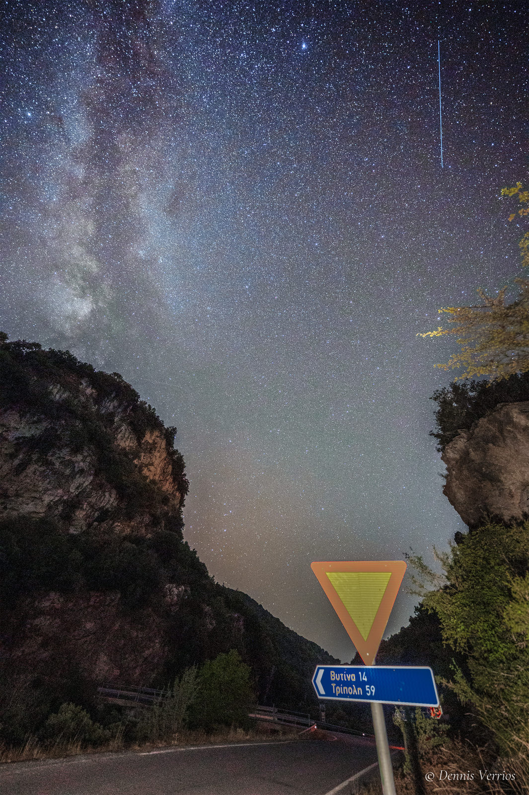 The Milky Way rising between rocky cliffs above a mountain road, with a yield sign and a road sign pointing to Vytina and Tripoli, in Arcadia, Greece. Photograph by Dennis Verrios.