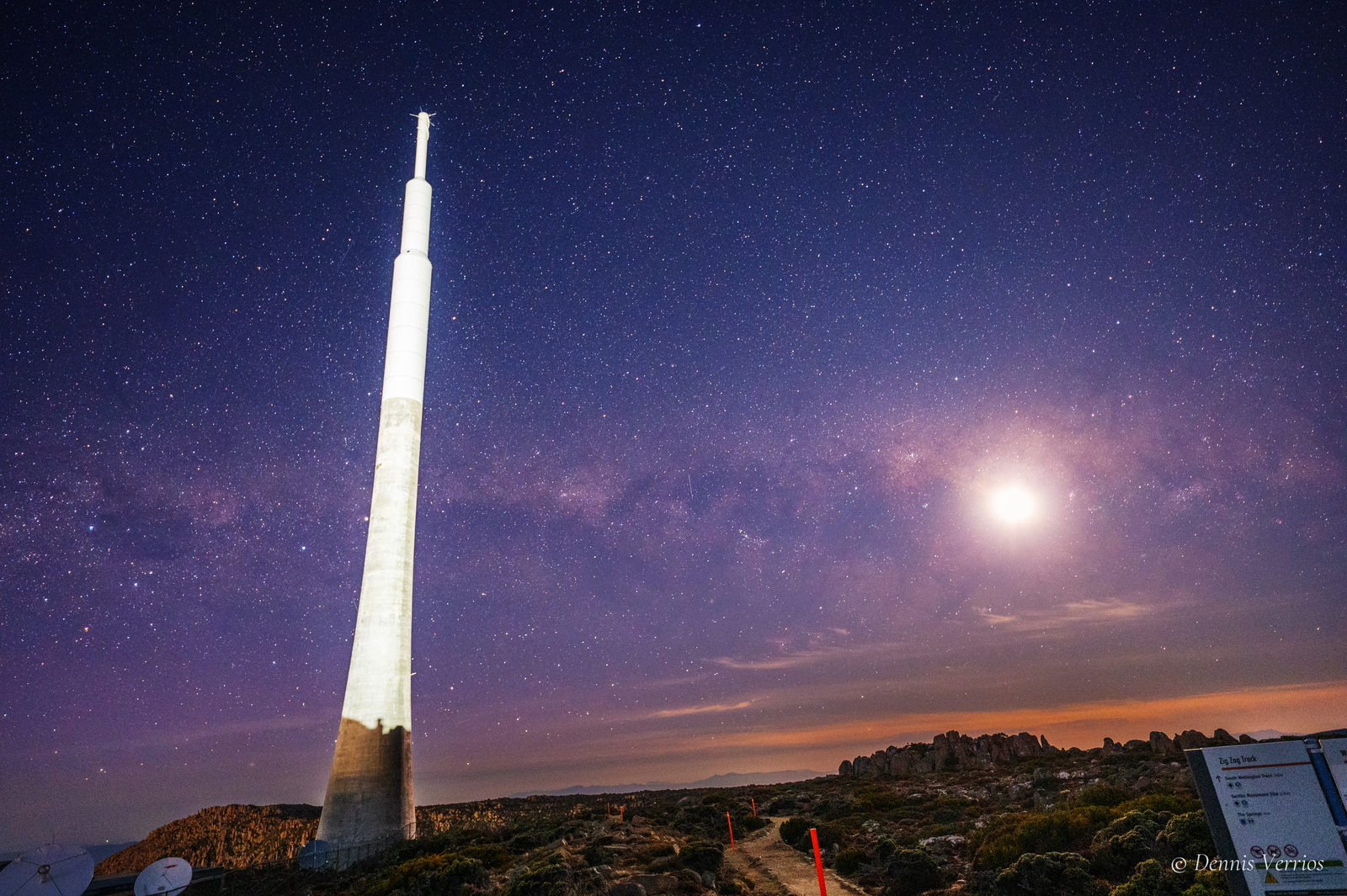 The tall white broadcast tower on kunanyi / Mount Wellington beneath a purple starry sky, with the bright Moon sitting in the Milky Way and the Zig Zag Track sign in the foreground. Photograph by Dennis Verrios.