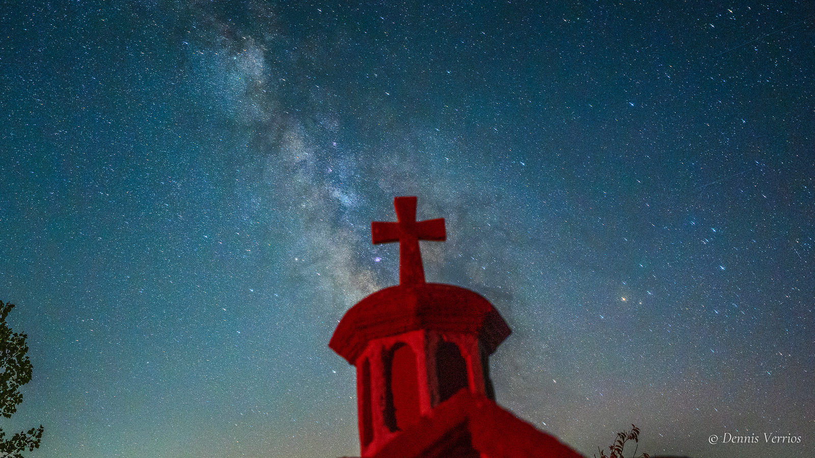 The cupola and cross of a tiny roadside church glowing bright red against a teal sky, with the Milky Way rising behind it, in Arcadia, Greece. Photograph by Dennis Verrios.