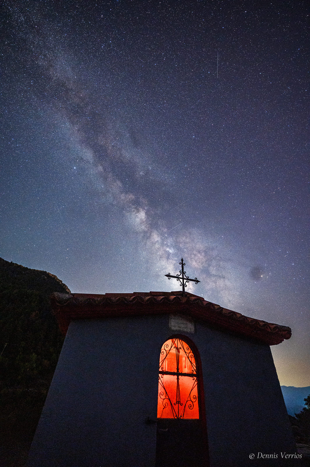 A tiny roadside church with a glowing red arched window and an iron cross on its roof, under the Milky Way near Dimitsana, Arcadia, Greece. Photograph by Dennis Verrios.