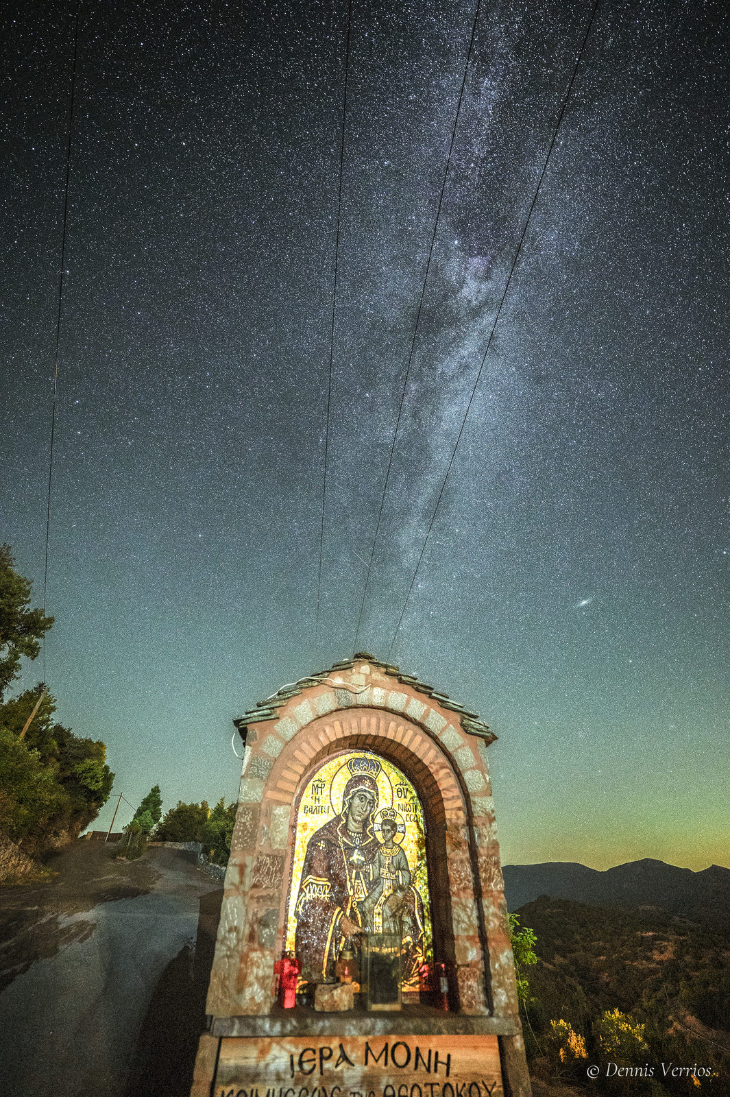 A stone roadside shrine holding a lit icon of the Virgin Mary, with the Milky Way streaming up into the sky above, near Valtesiniko, Arcadia, Greece. Photograph by Dennis Verrios.
