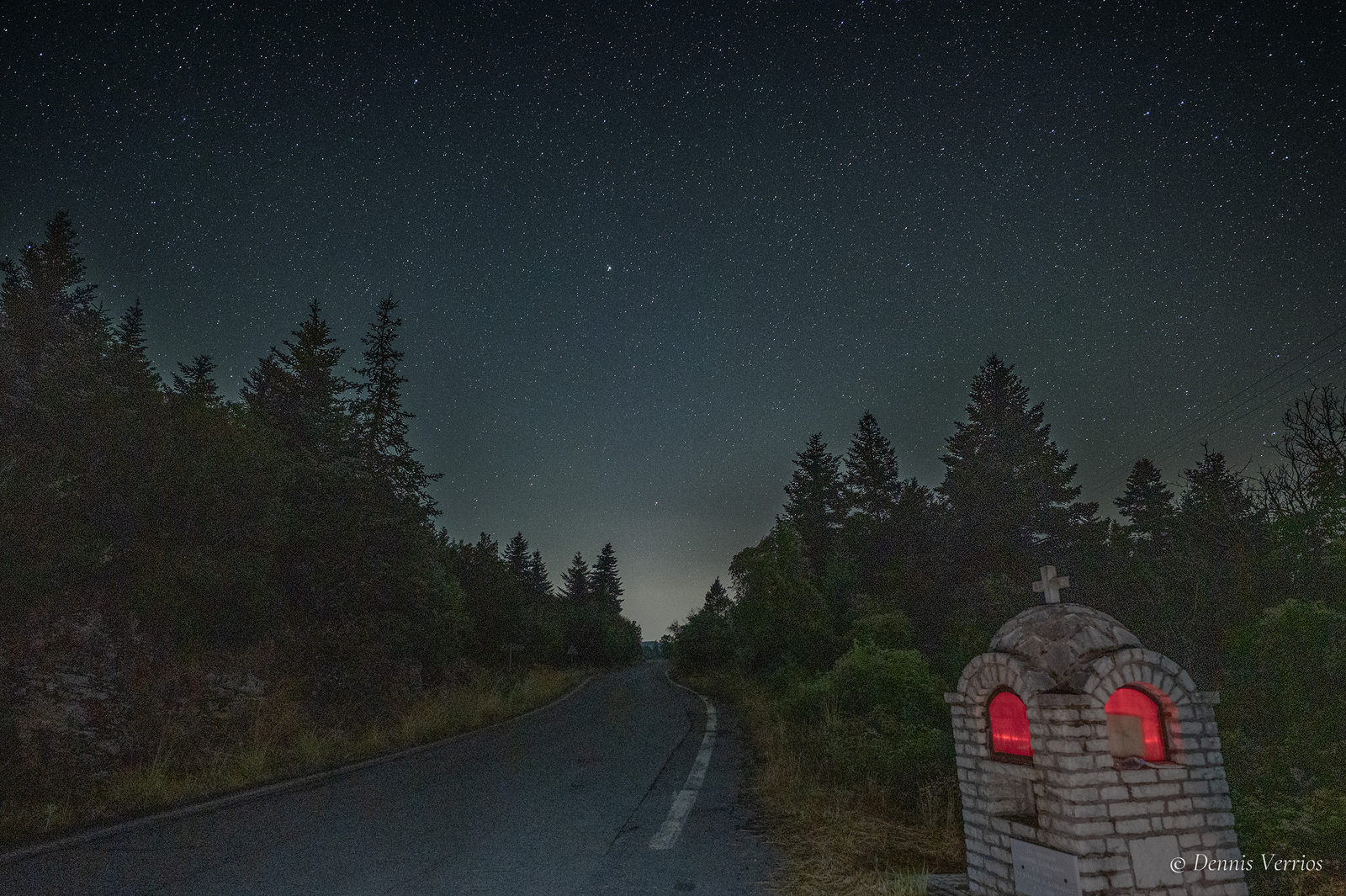 A small stone roadside shrine with glowing red windows beside a mountain road through fir trees, under a starry sky near Alonistaina, Arcadia, Greece. Photograph by Dennis Verrios.